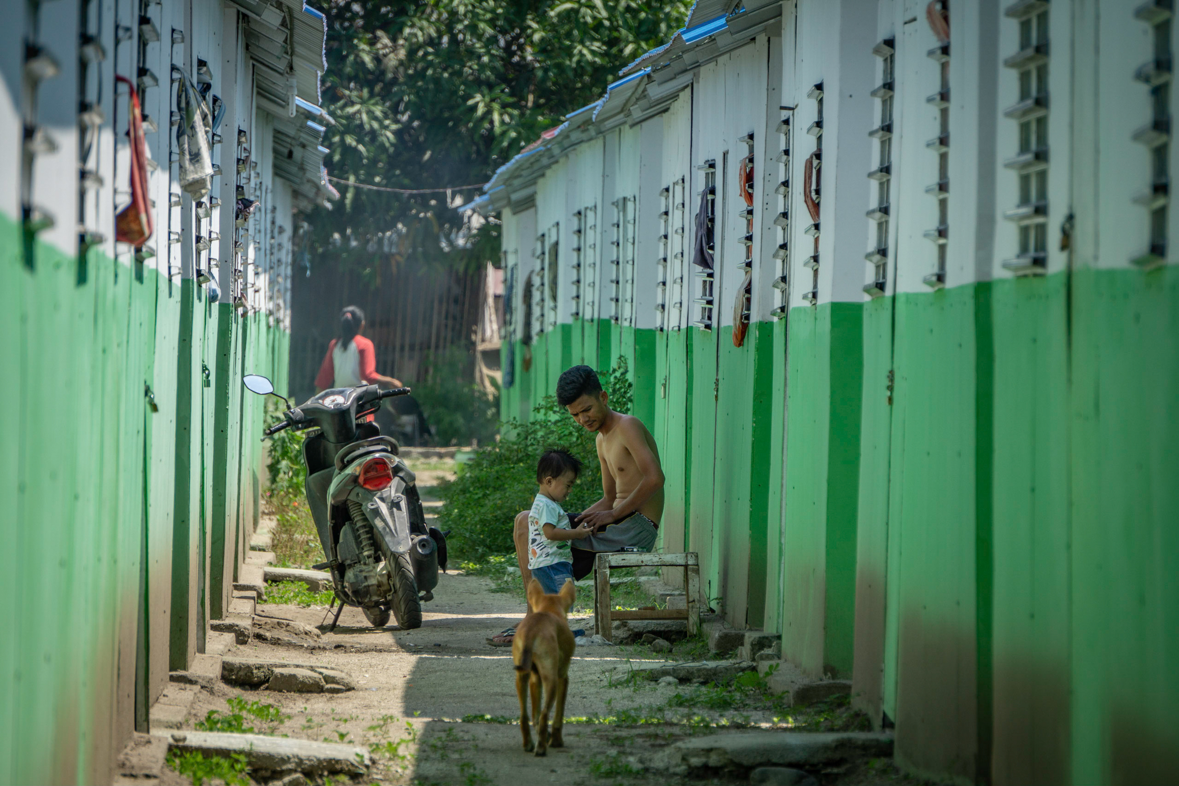 Photo of a man, child, dog, and scooter in a narrow passage between shelters.