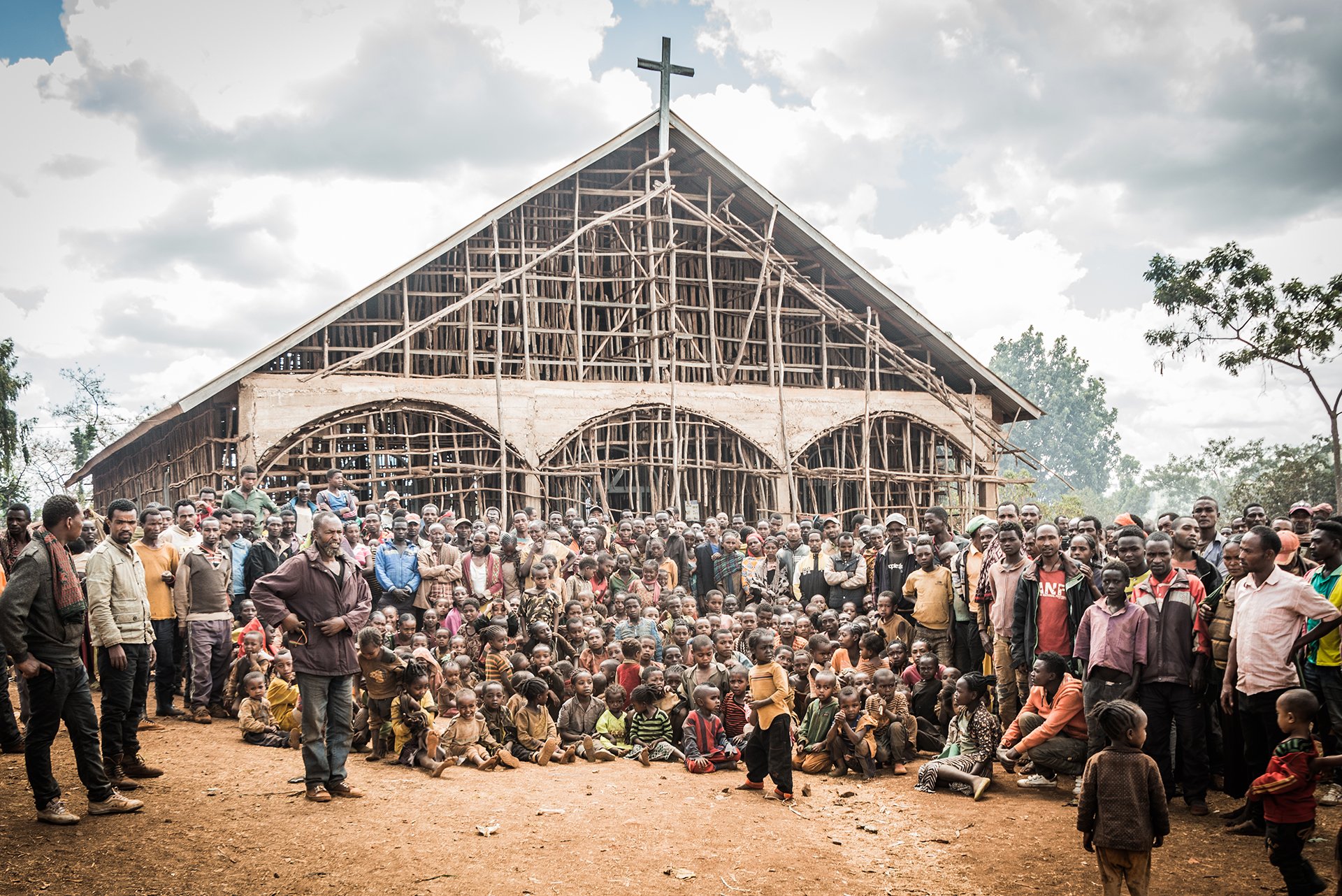 A large group of people standing for a portrait in front of a large, makeshift church against a dramatic sky