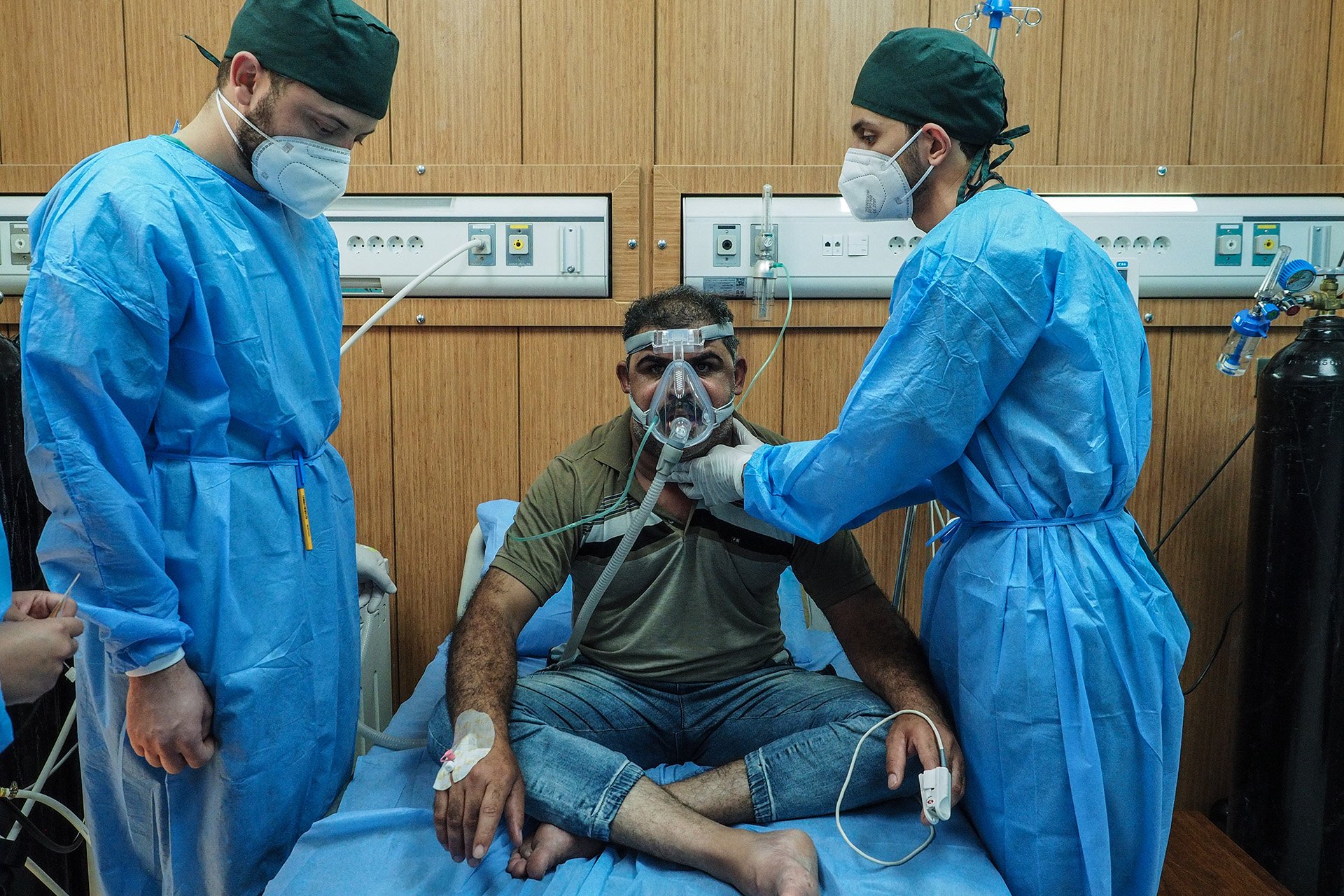 A man sits on a hospital bed while two people in blue medical scrubs assist him.