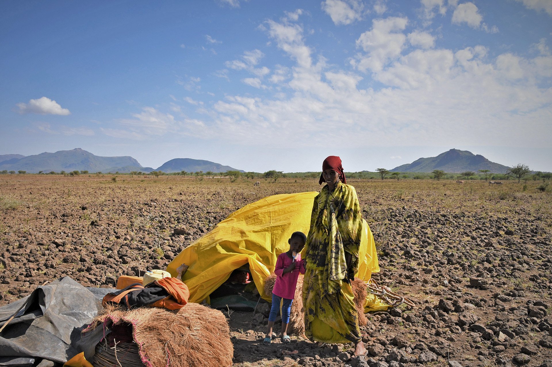 A woman and a small child stand next to a tent on an arid plain.