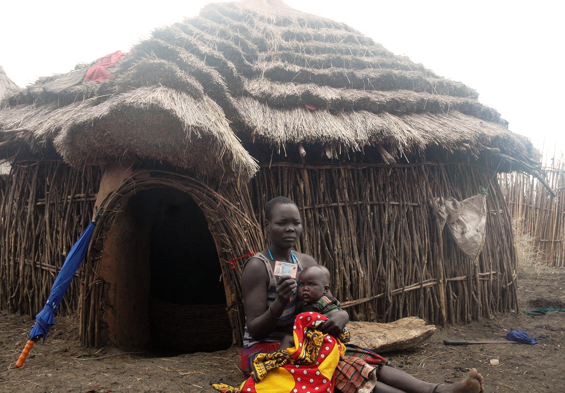 Photo of a woman with a chiild in her arm sits in front of a thatched hut in Uganda showing an ID card