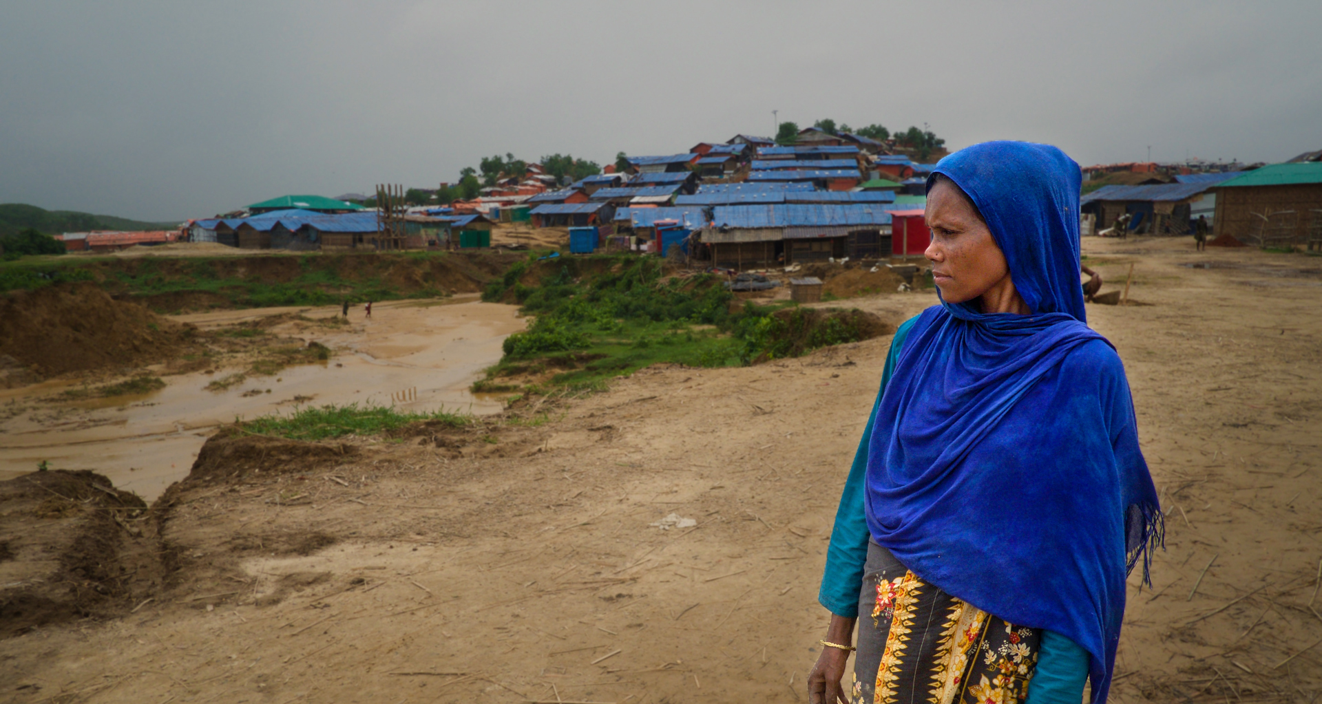 A woman in blue stands in front of a refugee camp looking to the side