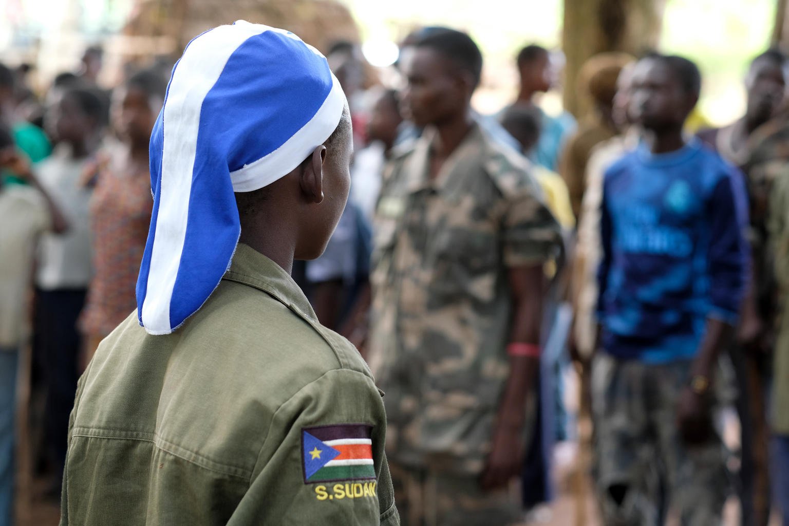 The back of a girl standing by a gun on the ground in front of ranks of other young people all facing away