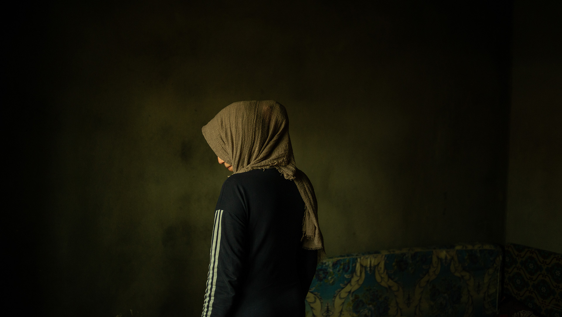 An image of a woman in a headscarf standing in a near empty room from behind, looking slightly down and to the side.