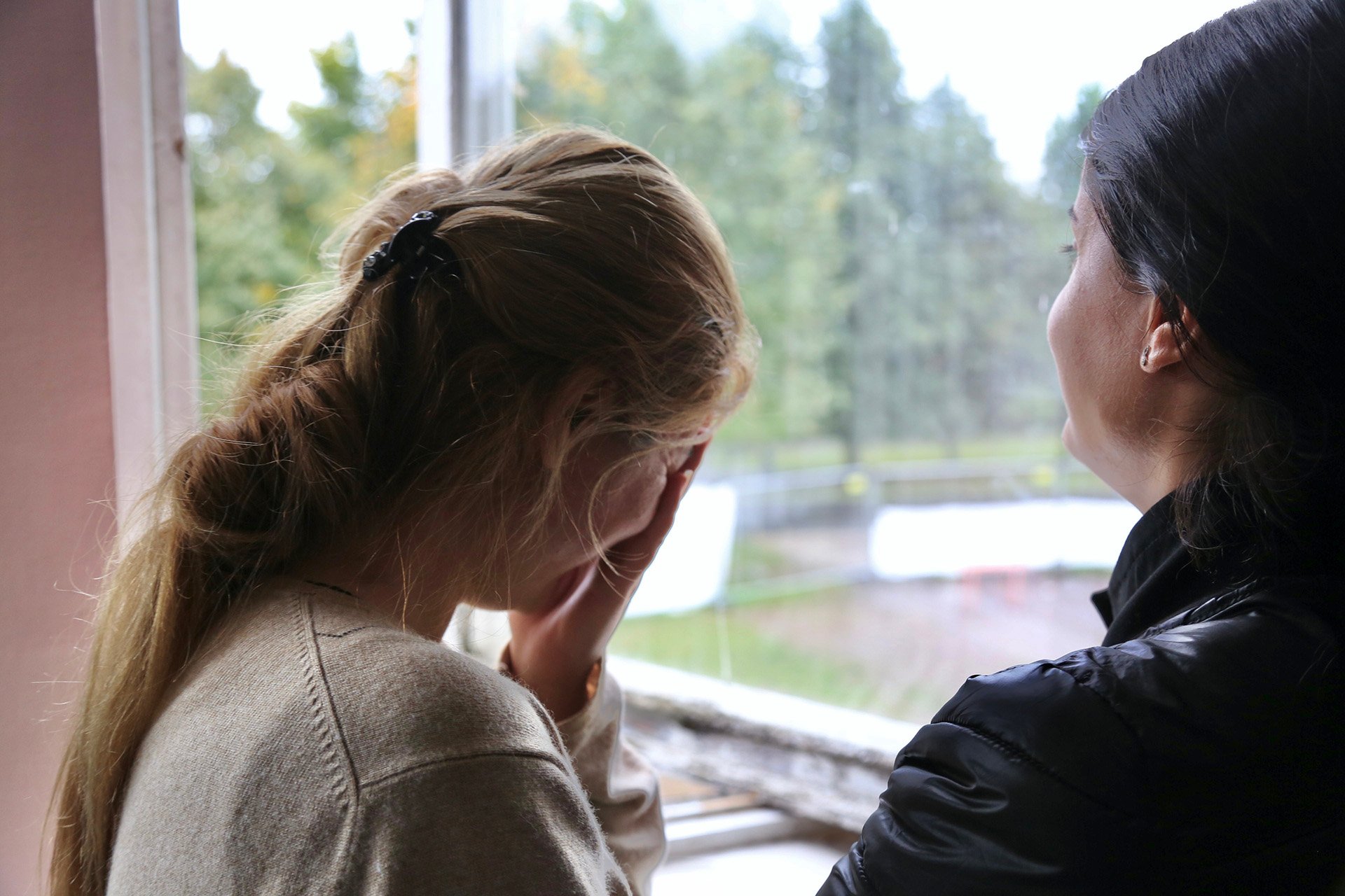 A woman stands facing a window, holding her head in her hands. 