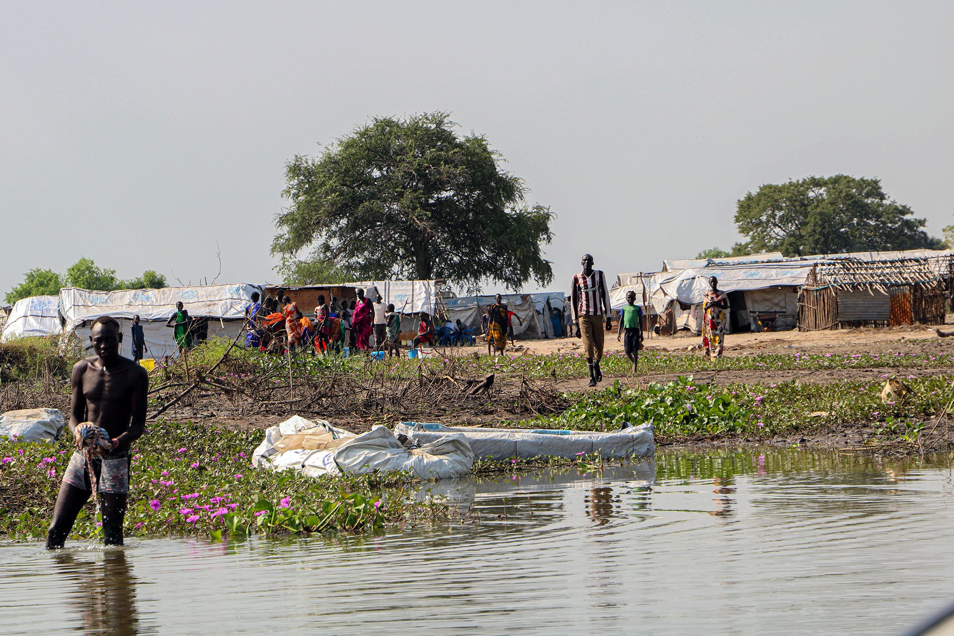 A man in the foreground of the photo washes clothes in the river. In the distance, low houses and other residents are visible. 