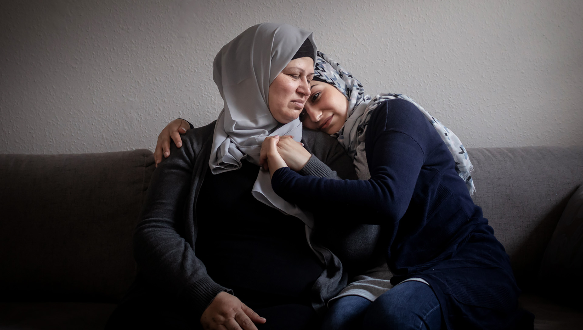 Two women sit on a couch and embrace. 