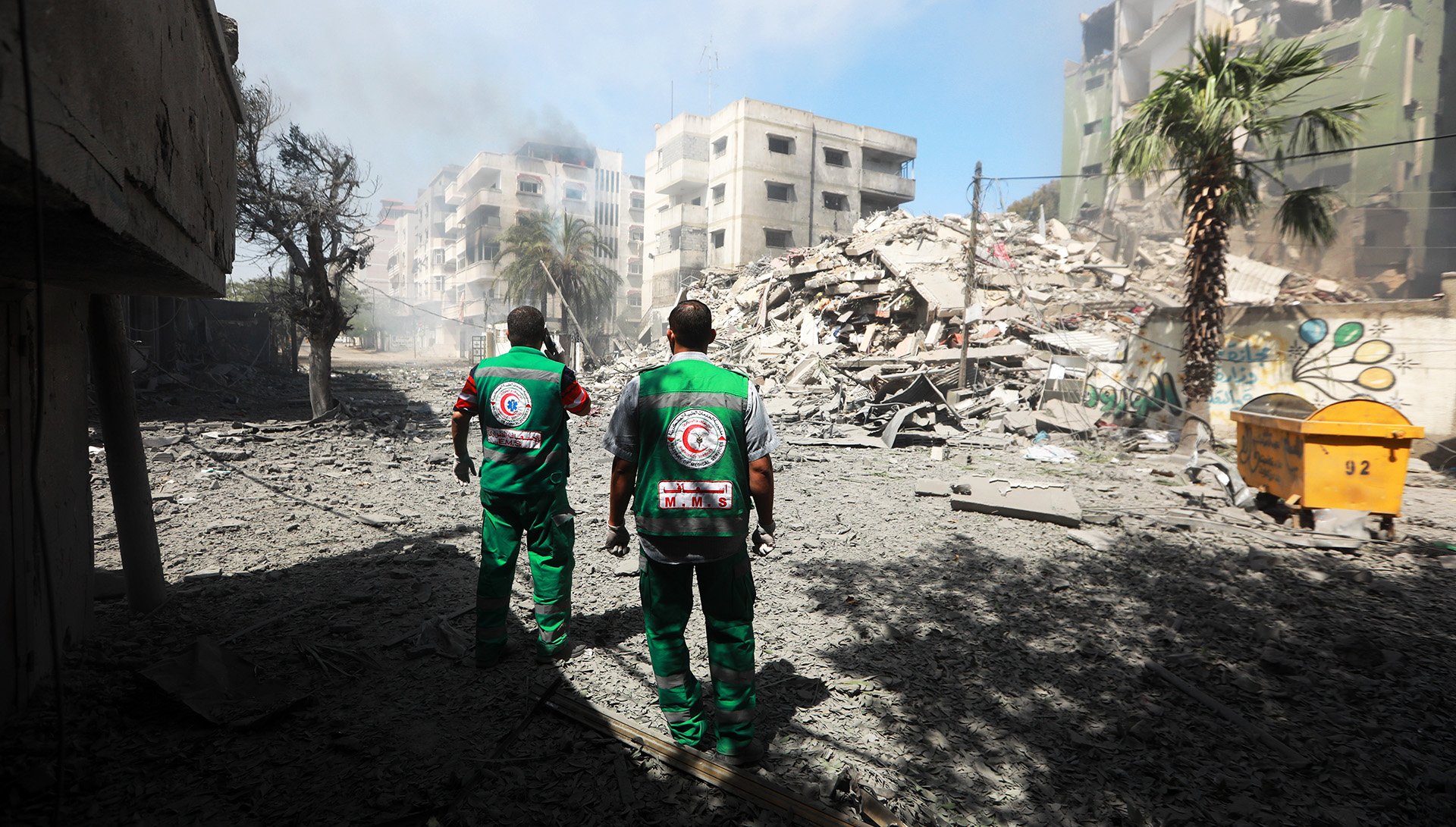 Two medics dressed in green uniforms stand in front of demolished buildings with their backs to the camera.