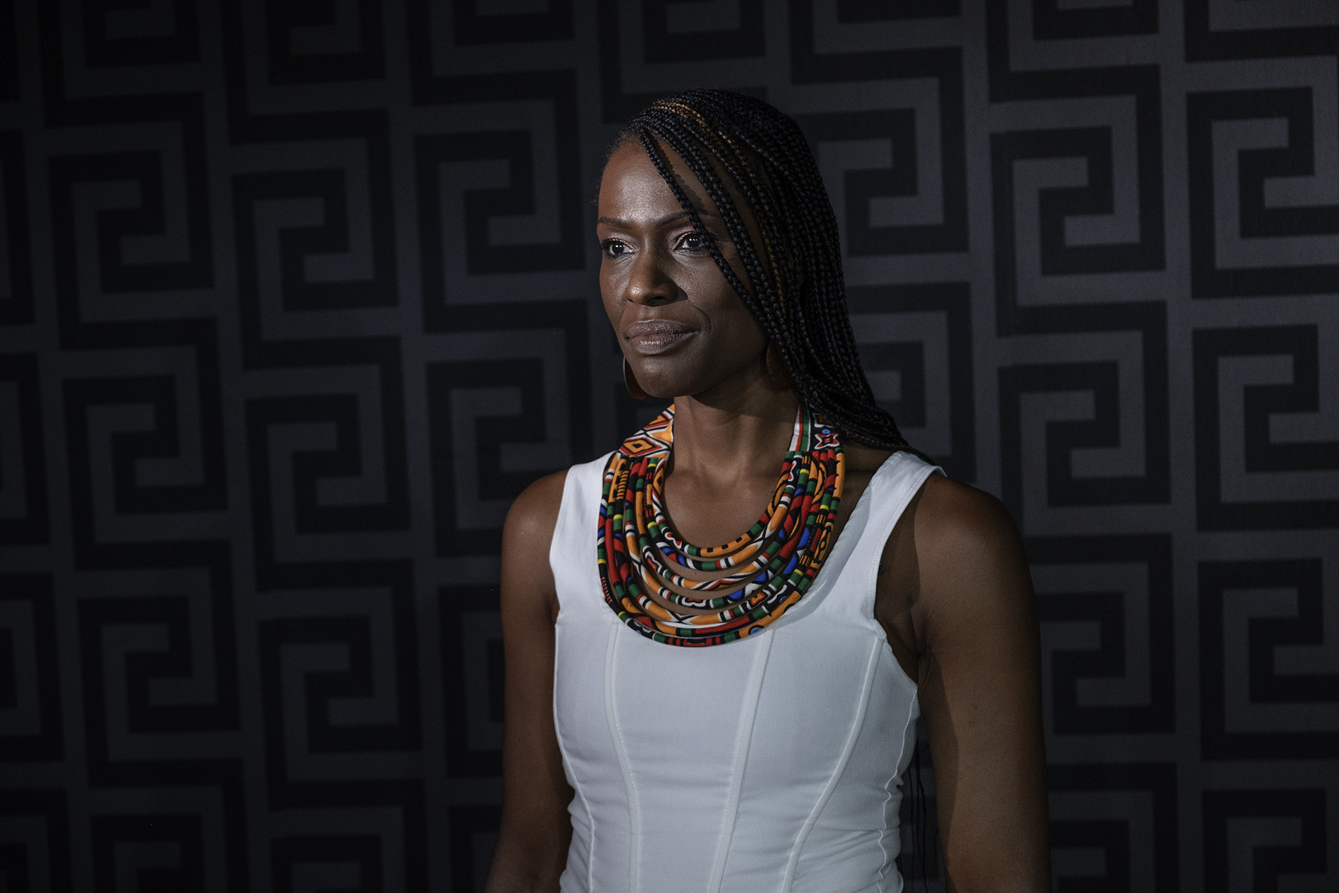 Portrait of a woman in a white dress and colourful necklace against a dark, patterned background.