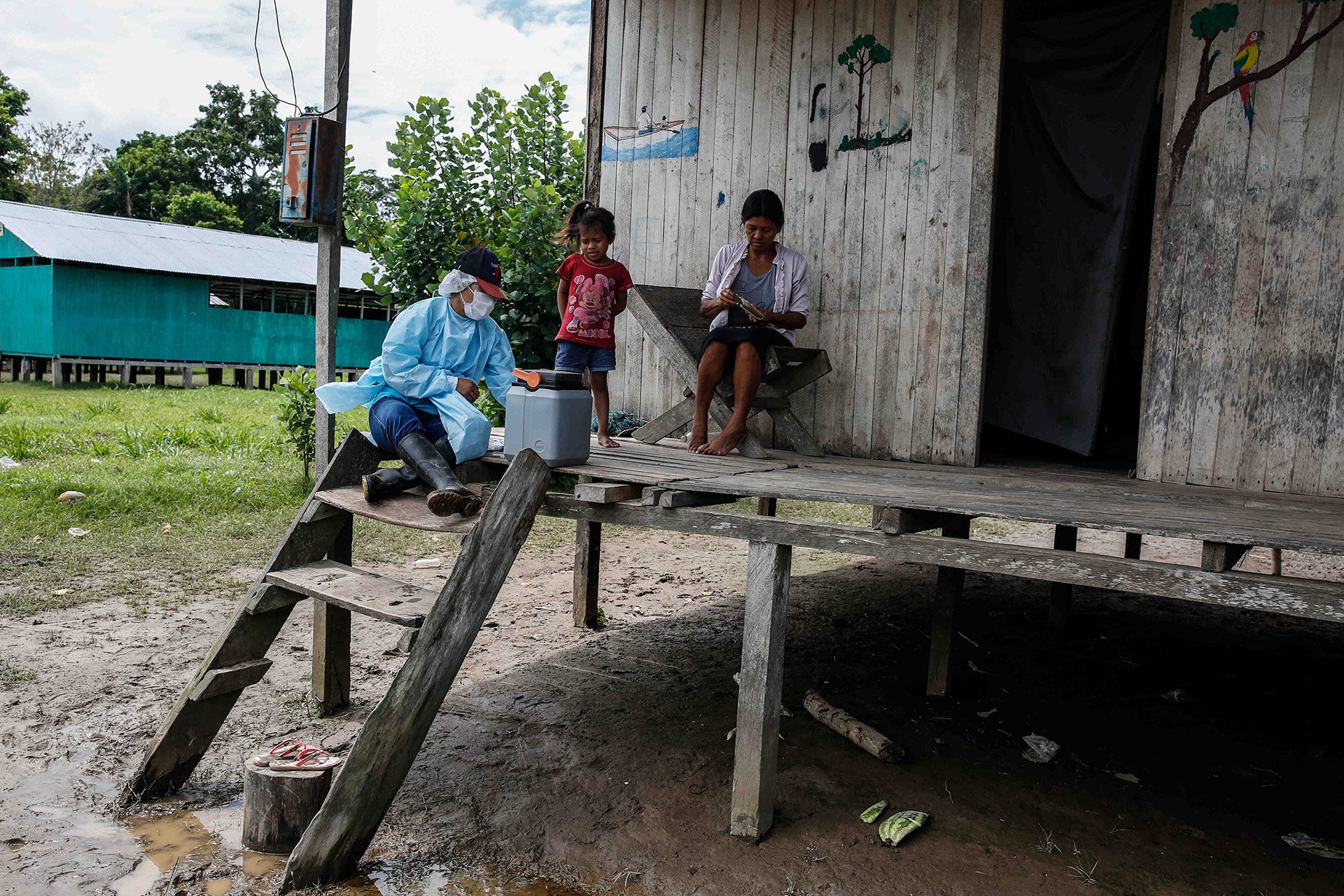 A nurse, woman, and young child stand on the porch of a building on stilts in a rural village.