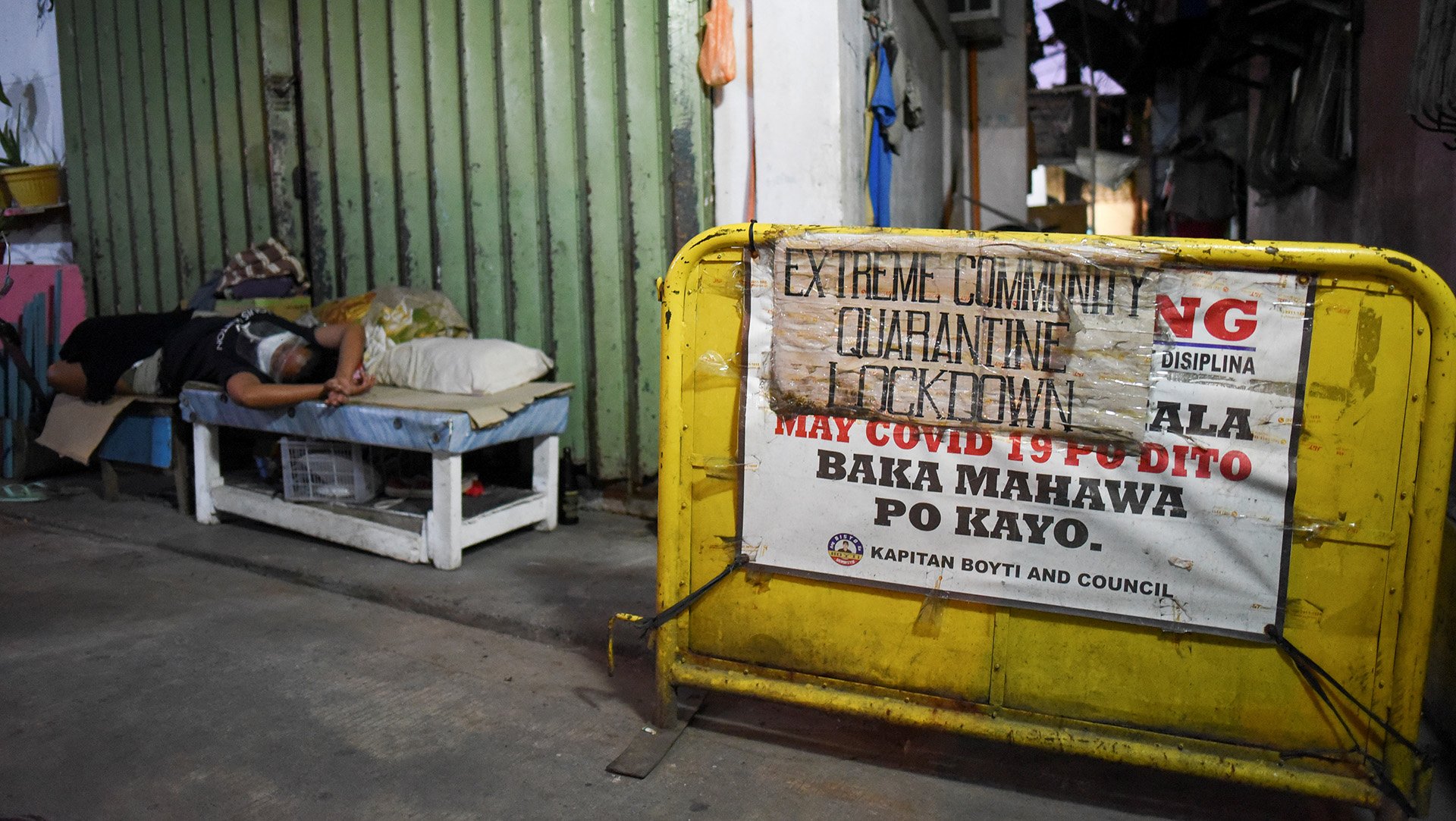 A sign blocks a city street indicating a local lockdown – in the background of the image, a figure lies sideways on a bench.