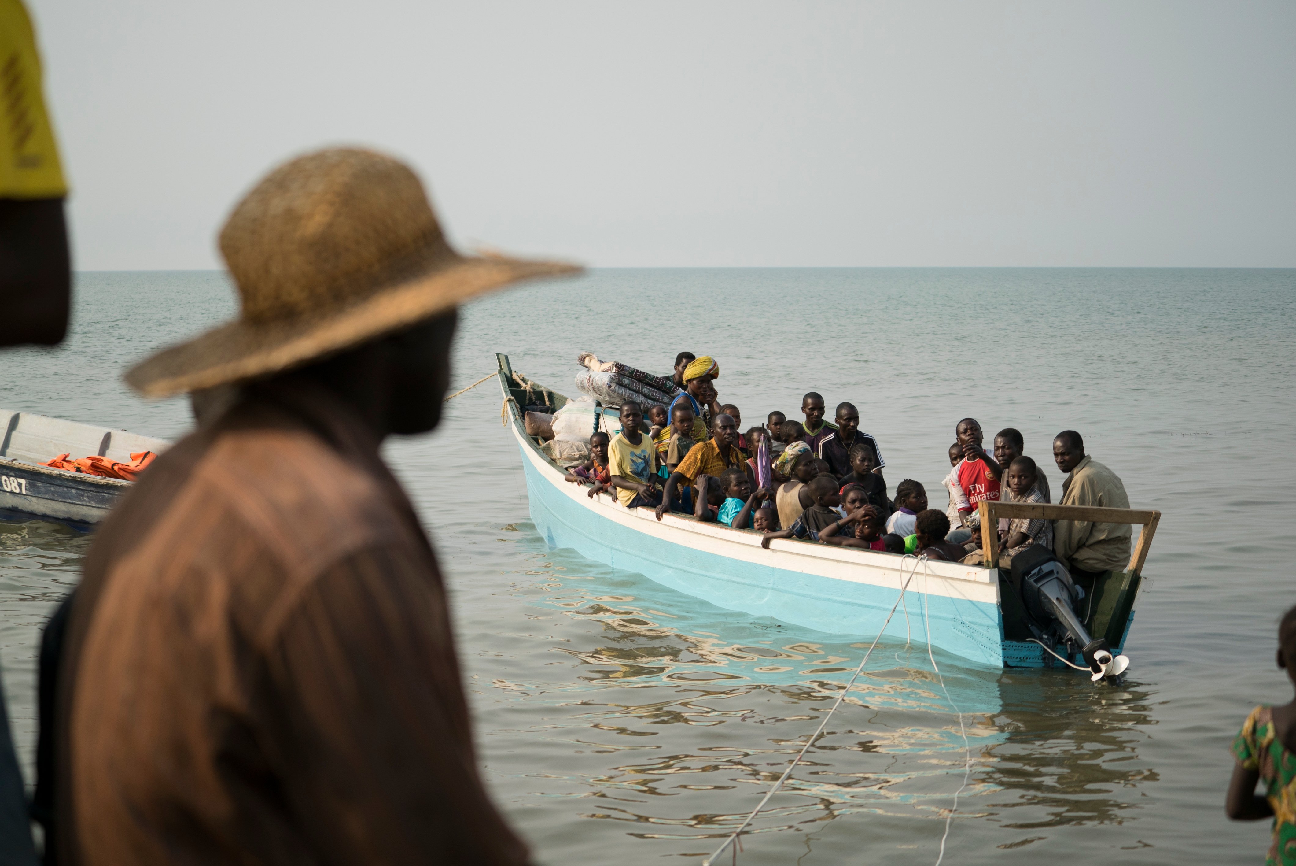 An out of focus man in the foreground looks out on a boat full of refugees from DRC landing in Uganda