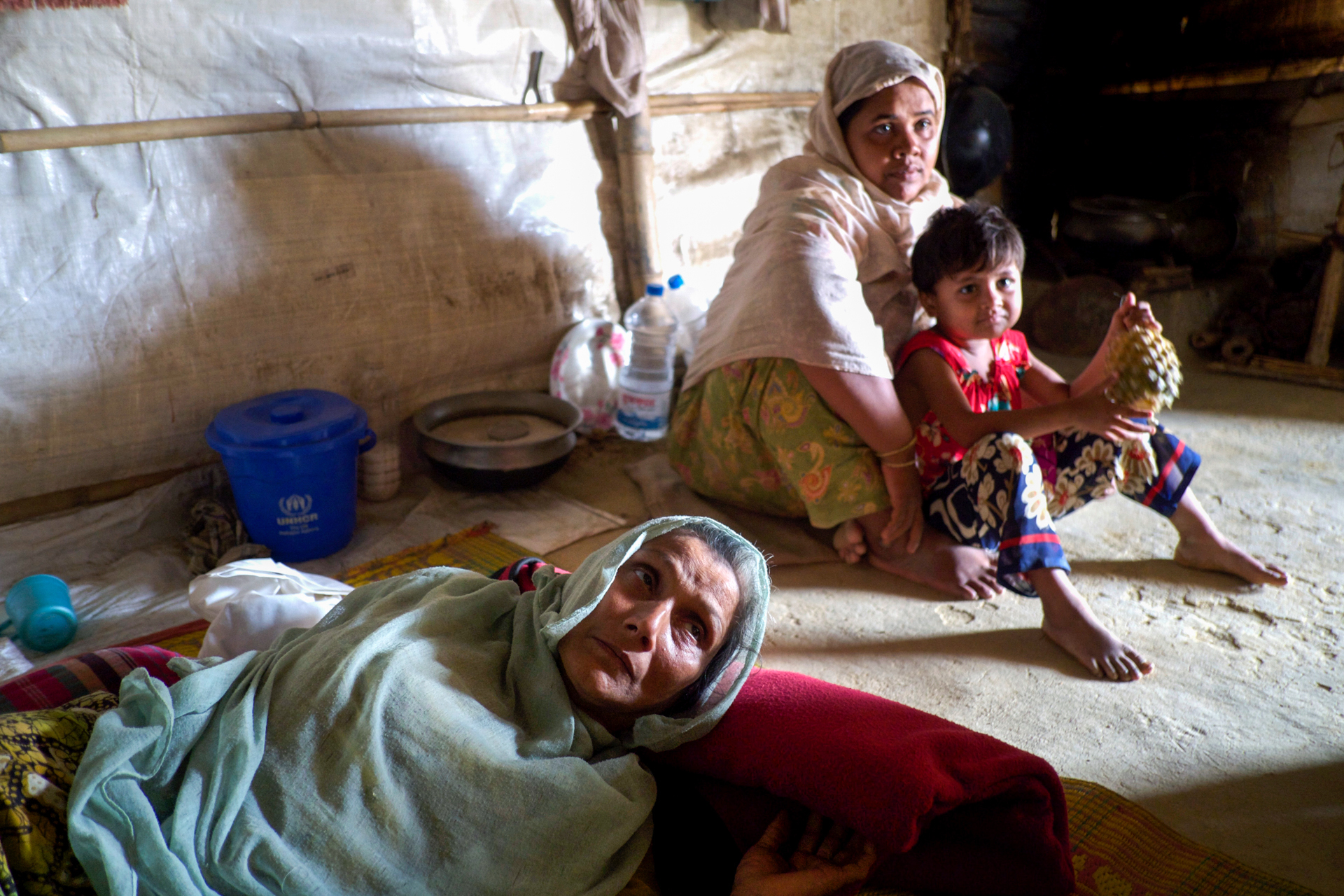 A woman in the foreground lays on her side while a younger woman and a child sit behind her on a dirt floor