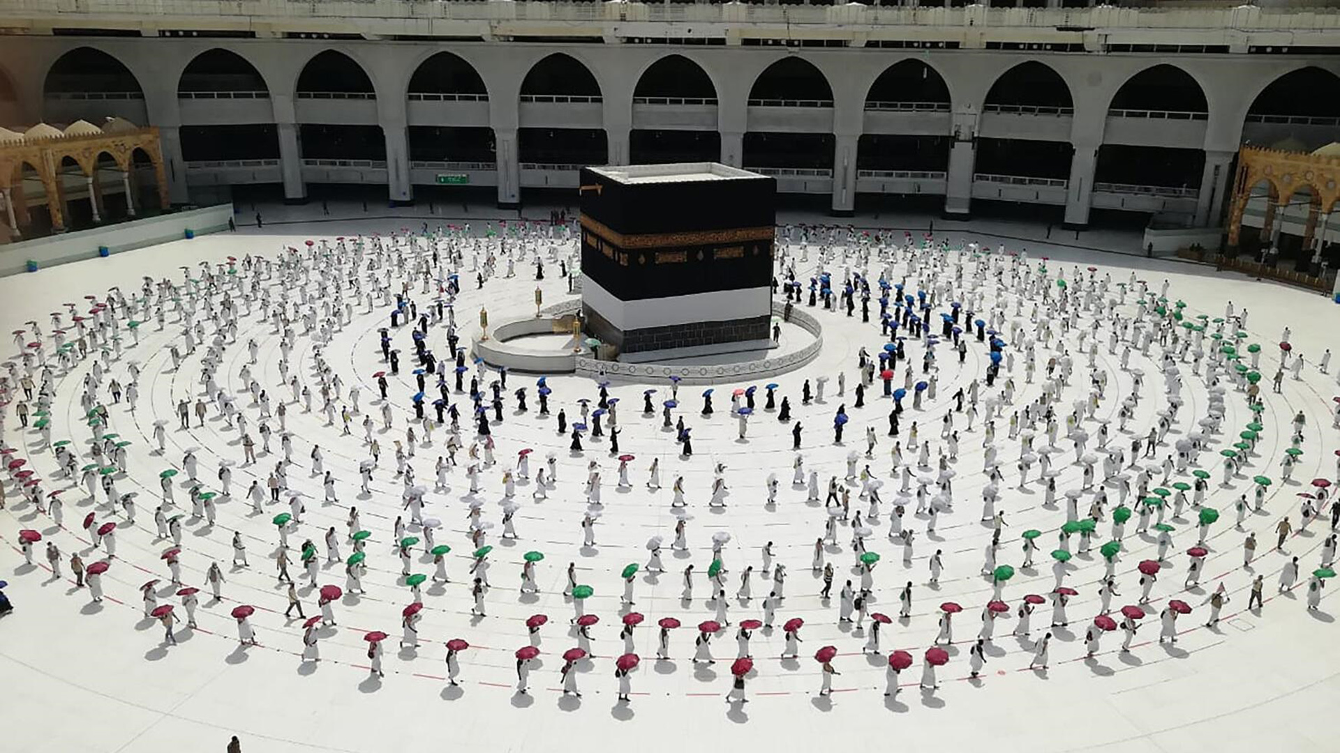 Sparsely populated Hajj ceremony with everyone circled, but spaced out and under colourful umbrellas.