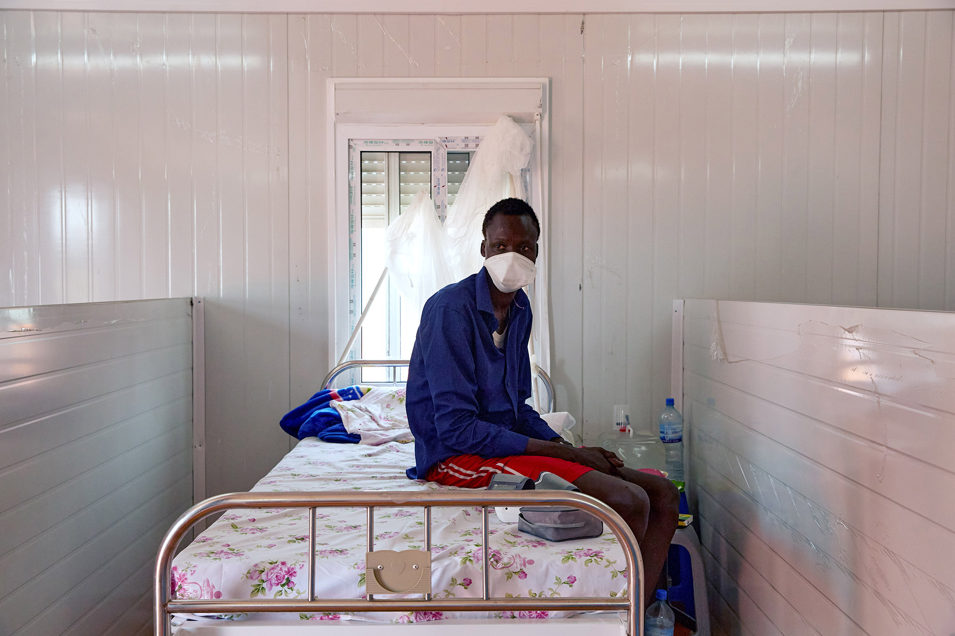 The image shows a young man sitting on a wrought iron hospital bed – a window half-covered by a curtain is above the bed