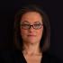A headshot of Sarah W. Spencer, a consultant and researcher specialising in technology, foreign policy, and aid. She is facing the camera face-front and has a closed-mouth smile. She wears glasses and a black blouse. The background is black.