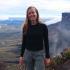 This is a photograph of Mie Dahl, an independent journalist covering politics, economics, conflict, and environment in Latin America, based in Bogotá, Colombia. She is pictured standing in nature with a view of a valley and some mountains on the right of the photo. She is wearing a black long sleeve shirt and grey hiking pants. She has long blonde hair and is smiling at the camera. 