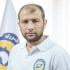 Headshot of Raed al-Saleh, the director of Syrian Civil Defence (The White Helmets). He is pictured wearing a polo shirt with the logo of his organization on the left. Behind him is a flag with the logo too.