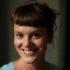 A headshot of Karolina Augustova. She's a Lecturer of Sociology and International Relations at Northumbria University. She's pictured smiling widely at the camera. Some shadows to the left of her face. She has brown hair that she wears up and has short blunt bangs. 