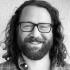 This is a black and white headshot of Sam Heller. He's pictured smiling at the camera. He wears dark-rimmed glasses and a plaid button-up shirt.