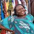 This is a headshot of Adior Ibrahim. She is the DEI program assistant at Cohere. She is pictued standing in front of different clothes, with her right hand on her hip and smiling at the camera. She is wearing a turquoise blouse.  