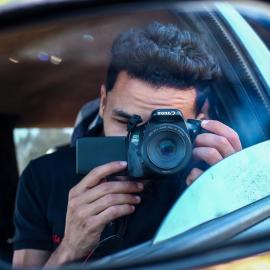 This is a headshot of Ameen Alokaley. He is a photographer from Derna, Libya. He is pictured taking a self-portrait. He is taking a picture of a side view mirror. His face is covered by the camera.