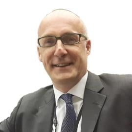A headshot of Oliver Lacey-Hall. He is pictured smiling wearing a grey suit and blue patternedtie.