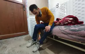A man sits at the edge of his bed, putting on his shoes. 