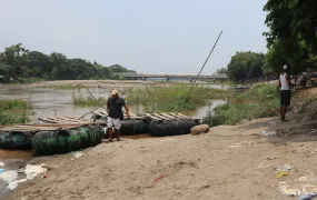 In the foreground we see two rafts using tires for floatation, a man walks between both of them. Behind is a river.