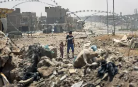 Two children walk amongst rubble as seen through a barbed wire fence