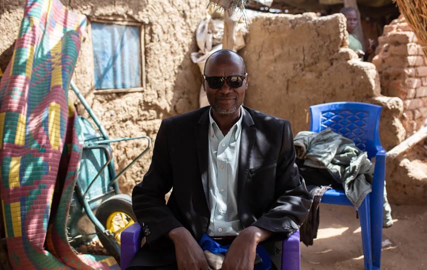 A man in sunglasses sits facing the camera in a suit.