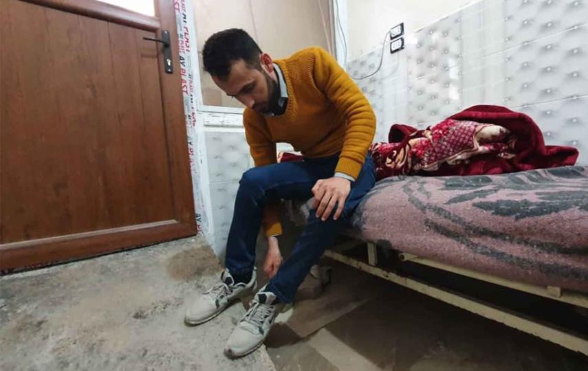 A man sits at the edge of his bed, putting on his shoes. 