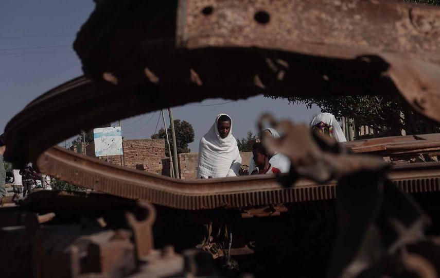 In the foreground are the scraps of a war tank, through a hole in the scraps we see a man dressed in white. His face is looking downwards.