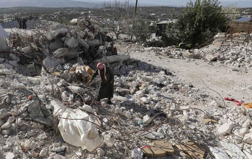 A man is standing on a pile of rubble. He is bent at the waist looking through the rubble. 
