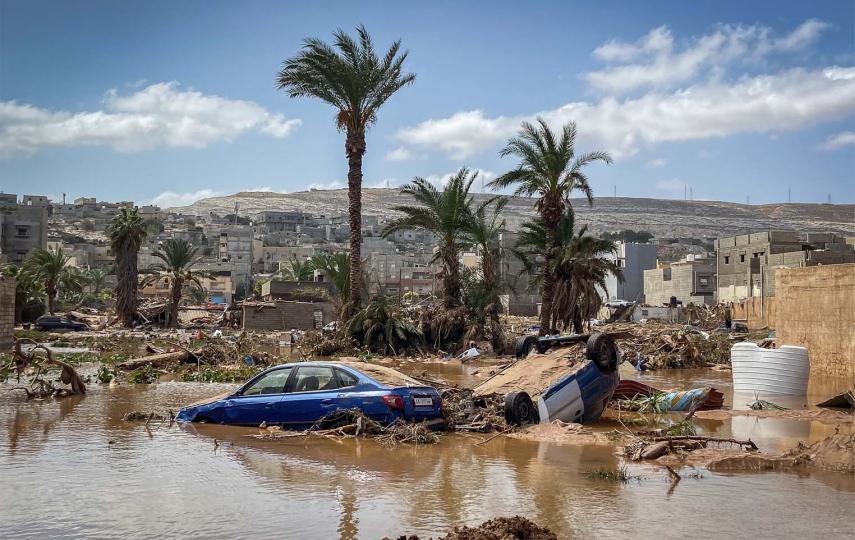 This photo shows the aftermath of the 2023 Libya flood. Pictured in the daytime, there are two cars at the centre of the frame. One is flipped over, mud can be seen on its undercarriage. Another car is next to it, on the left. It’s blue and its wheels are submerged in water. The majority of the lower half of the photo shows water. You can see damaged buildings around and a few palm trees behind the cars.