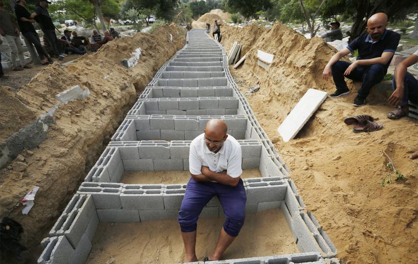 A man is sitting on cinder blocks that have been placed around graves that have been dug up on the ground. His hair is tilted downwards and his arms are crossed over his stomach. Behind him we see rows and rows of graves that are prepared to bury Palestinians killed in Israeli airstrikes at a cemetery in Deir al-Balah, a city in central Gaza, on 17 October.