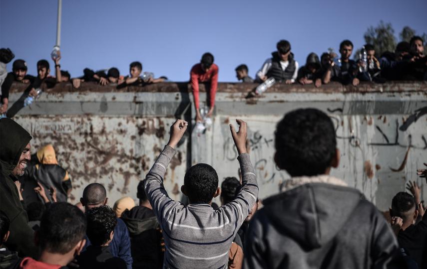 This is a medium shot showing a group of Palestinians flock to the truck carrying drinkable water as they face the threat of hunger and thirst in Rafah, Gaza on December 11, 2023. At the centre we see the back of a young person with their hands raised.