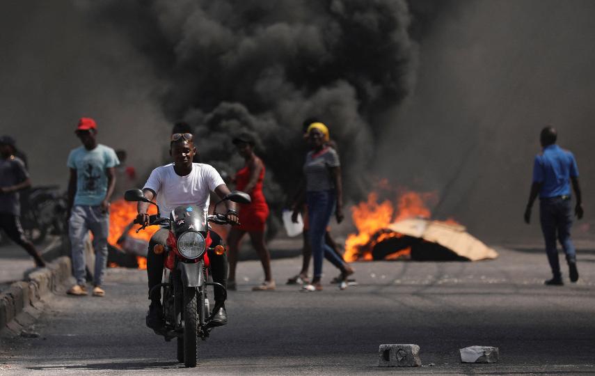 A man drives past a burning barricade in a motorcycle.