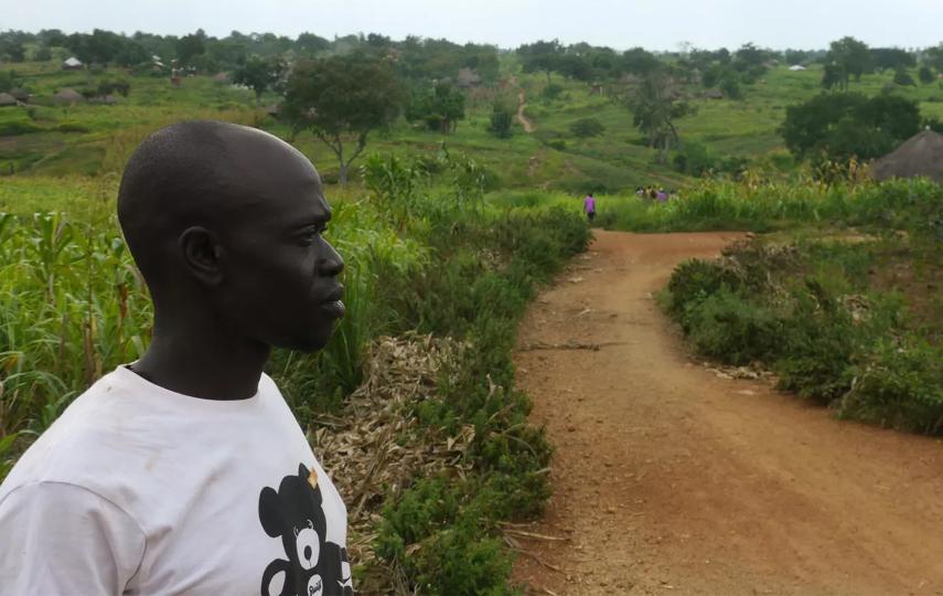 Isaac Ankora is pictured at the left side of the frame, looking into the distance towards the right. A dirt road surrounded by green foliage is in the background.