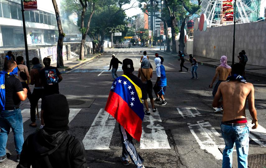 A picture of the street in Venezuela with people protesting. In the middle of the frame we see a person walking with a Venezuelan flag draped over their back.