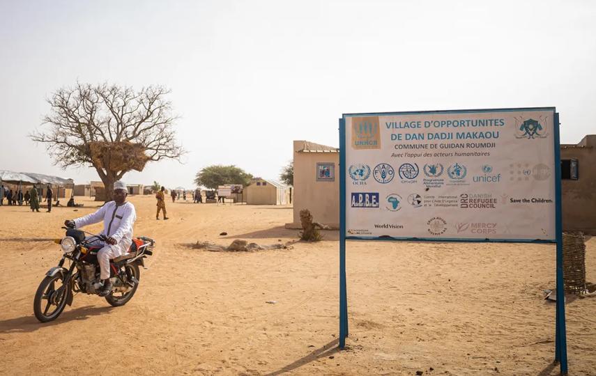 A landscape, wide-angle shot, showing a man in a scooter riding past a sign that shows this is an Opportunity Village in Dan Dadji, Niger.