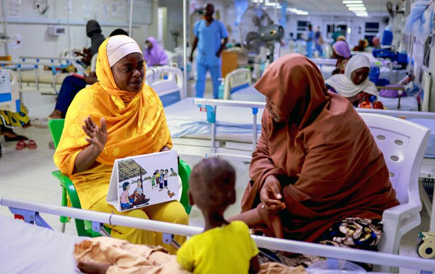 In a hospital, two women sit next to each other by the hospital bed where a young child sits as well. We only see the back of the child while we see the two woman engaging in an interaction.