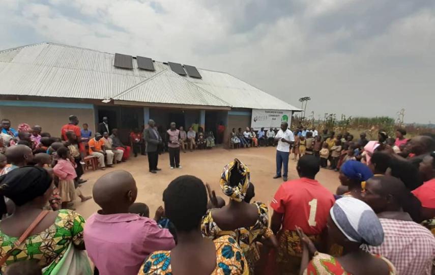 An outdoor meeting with people sitting and standing in a circle around a few people presenting.