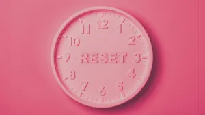 A round wall clock entirely in shades of pink, mounted on a pink background. The clock has raised hour numbers from 1 to 12 around its face but no hour or minute hands. At the center, where the clock hands would normally be, the word "RESET" is prominently embossed in uppercase letters, replacing the hands.