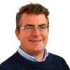A headshot of Ian Scoones. He is the Principal Investigator of Pastoralism, Uncertainty and Resilience (PASTRES) Programme, Institute of Development Studies, University of Sussex, UK. He is photographed against a white background. He's wearing a deep blue sweater and a light blue button-up shirt. He's also wearing glasses and smiling at the camera.