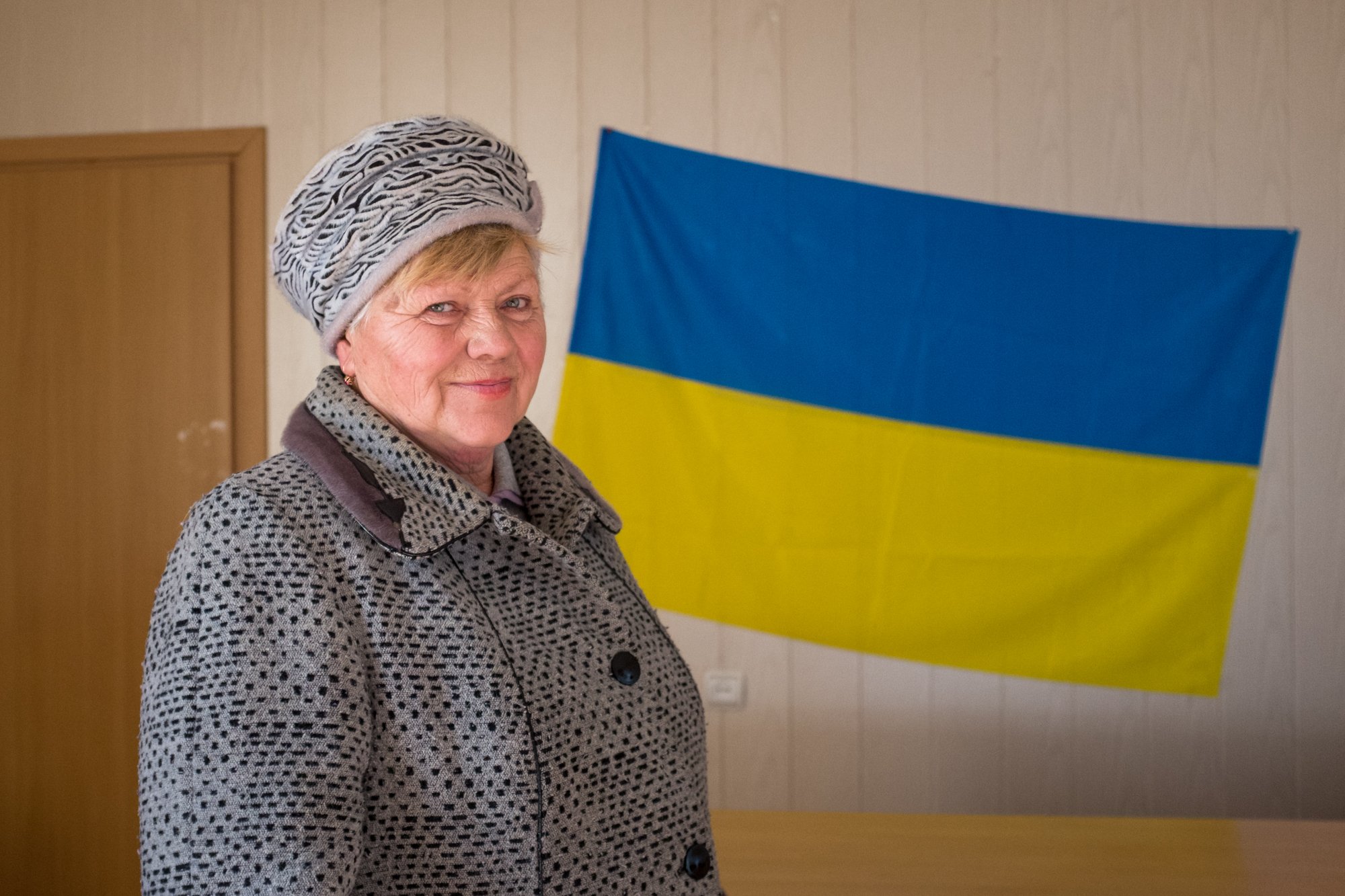 Photo of Tamara, head of a club for children in Pavlopil, in front of a blue and yellow flag indoors in Ukraine