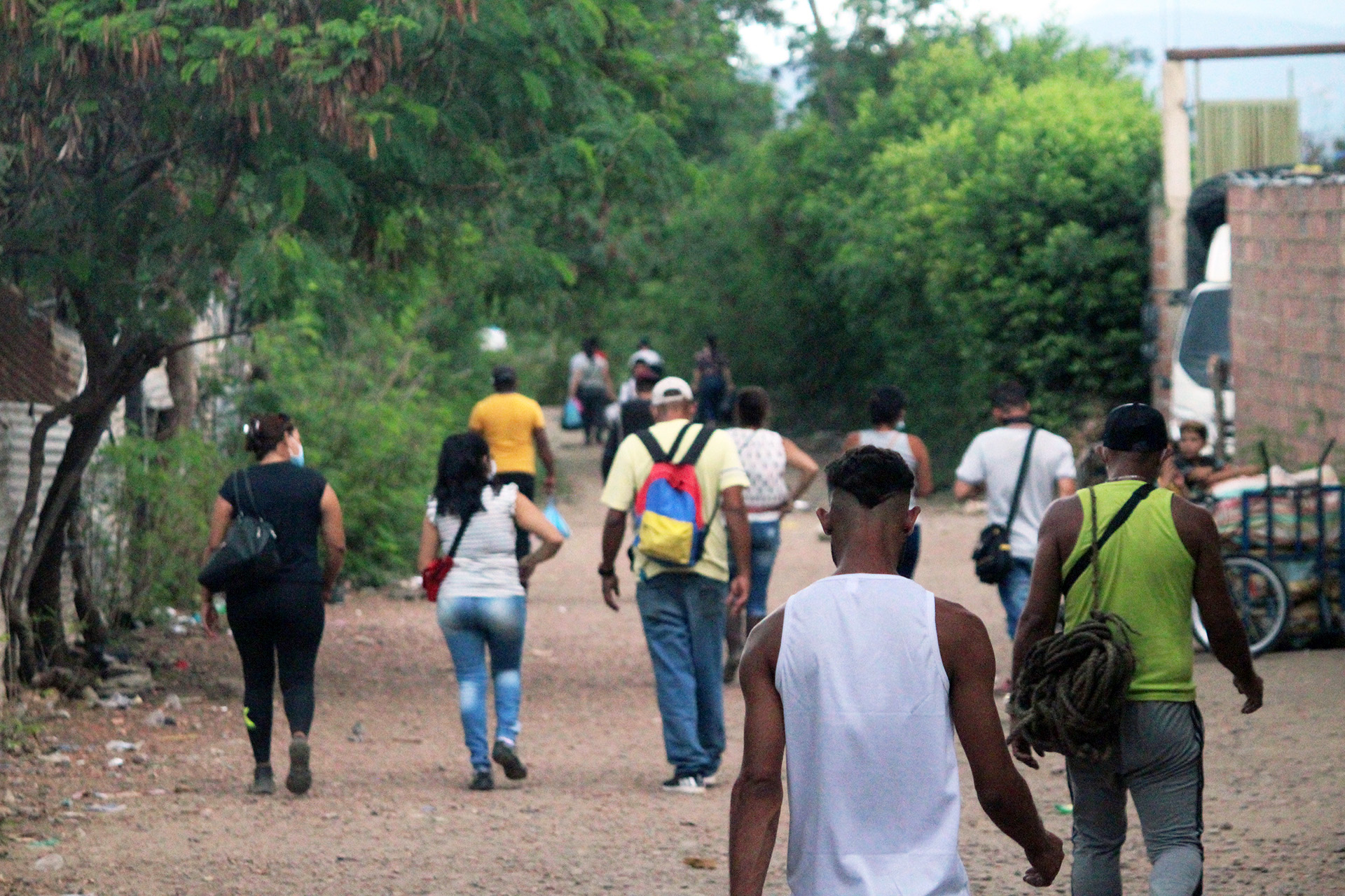 A group of migrants walk along a dirt road lined by greenery with their backs to the camera. 