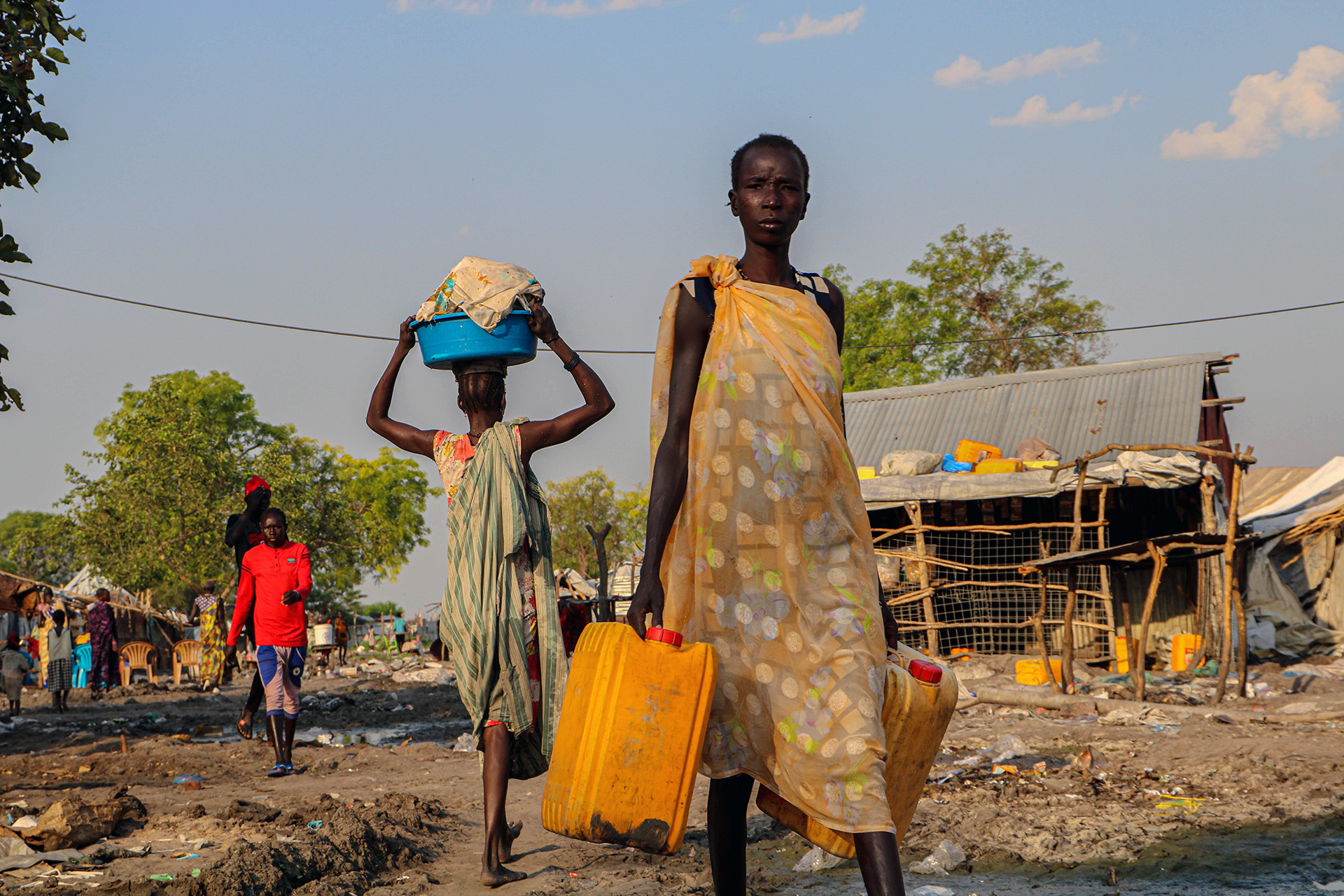 A woman walks towards the camera carrying cans of water.