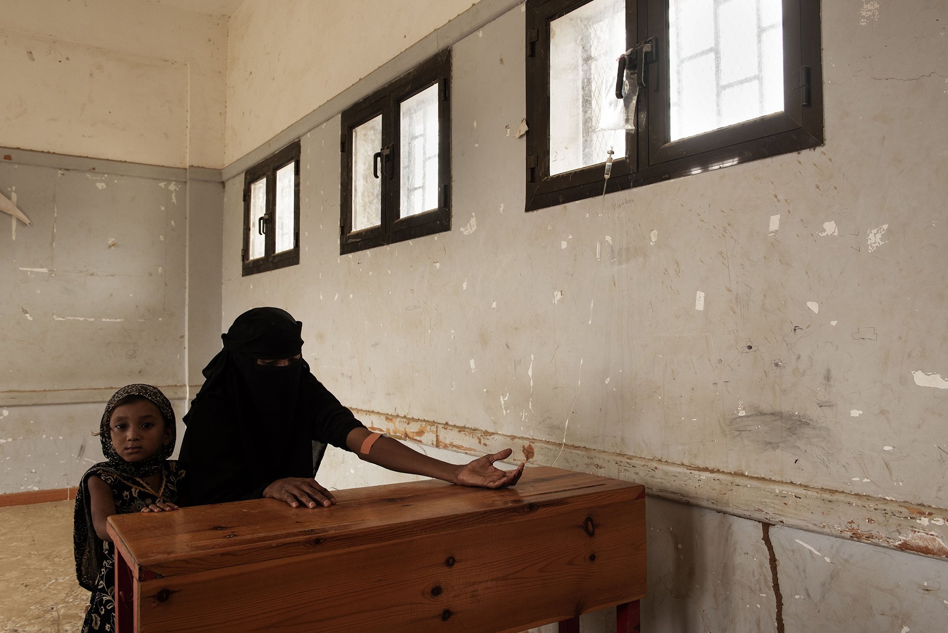 A woman sits at a table in an empty room with a young girl, the woman is hooked up to a drip hanging from a window.