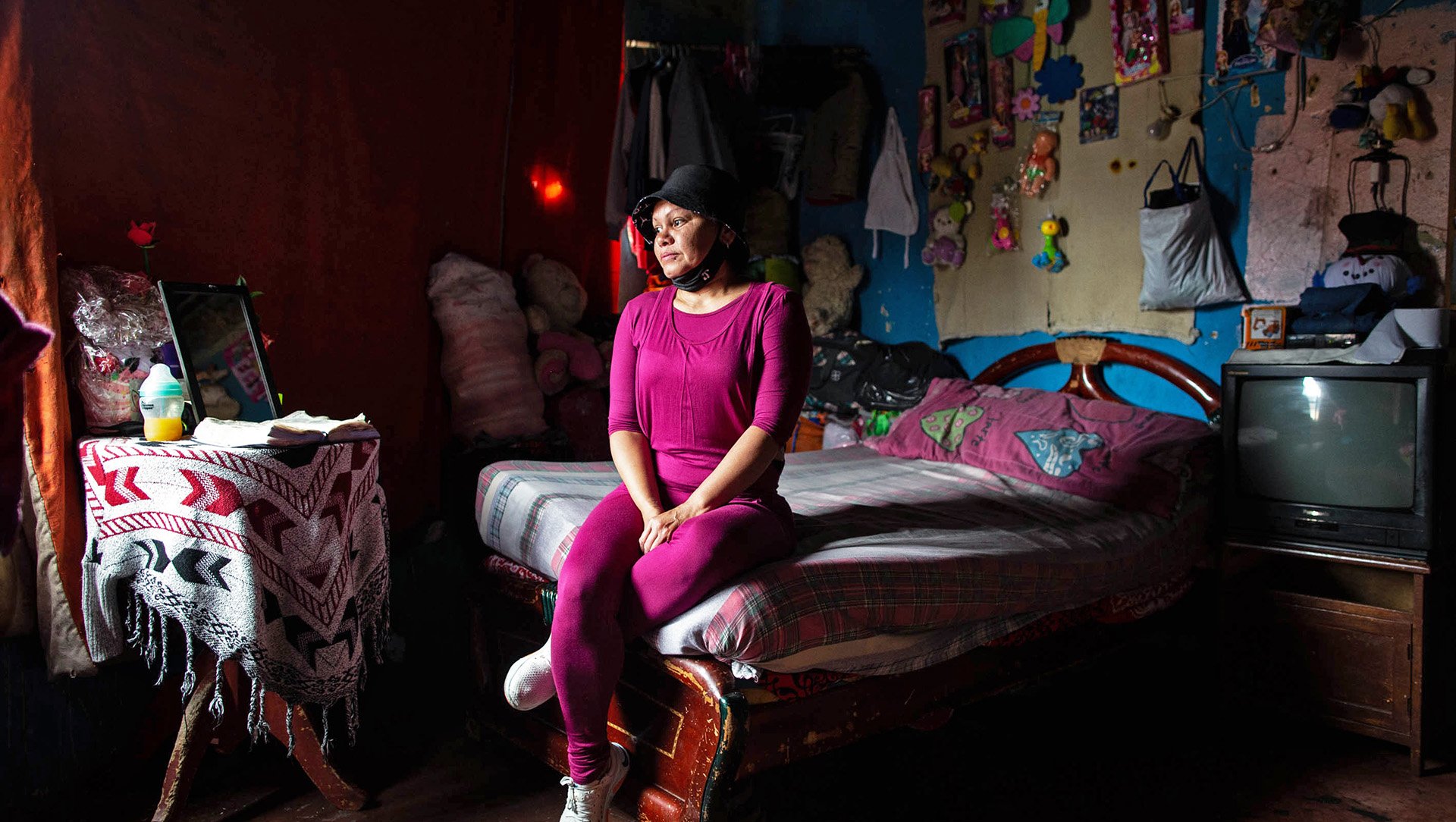 A woman sits on the edge of a bed in a darkened bedroom, gazing down at the floor to the right of her.